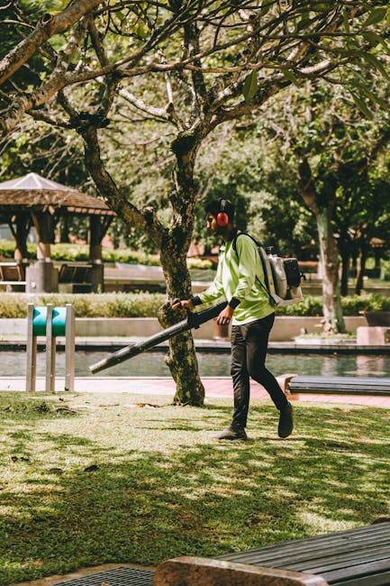 A person in a bright green jacket, black trousers, and wearing red ear protection is using a leaf blower to clear grass and fallen leaves under a large, mature tree with textured, moss-covered branche
